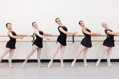 Adult Ballet Advanced Ages 18+ SUMMER - Hanover Theatre and Conservatory Five ballet dancers in black leotards and pink tights stand at a wooden barre in a bright studio, poised in a line.