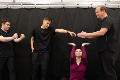 Adult Improv Ages 18+ - Hanover Theatre and Conservatory Four adults in black clothing perform a coordinated pose against a black backdrop; a woman kneels with arms raised while three men extend their arms toward her.