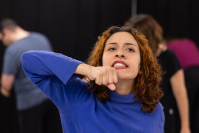 Adult Intro to Character and Clown Ages 18+ - Hanover Theatre and Conservatory Woman with curly red hair in a blue top extends her arm in a punch gesture toward the camera, likely in rehearsal or dance.