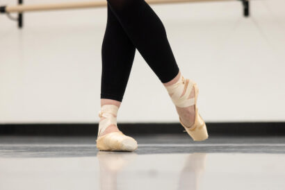Adult Pointe Ages 18+ SUMMER - Hanover Theatre and Conservatory Ballet dancer en pointe with ribboned ankles and crossed legs, in a studio with a barre in the background.