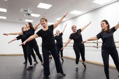 Adult Tap 1 Ages 18+ SUMMER - Hanover Theatre and Conservatory Group of older adults in black workout clothes performing a dance move with arms raised in a studio with a barre along the wall