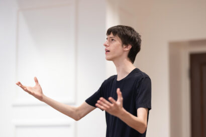 Audition Singing for Teens Grades 9-12 - Hanover Theatre and Conservatory Young man in a navy T-shirt speaks and gestures with outstretched arms in a bright room