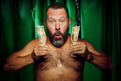 Bert26_1920 x 1080 hero image - DCM - video card - FB event - Hanover Theatre and Conservatory A shirtless man holds two overflowing glasses of champagne and looks excitedly at the camera