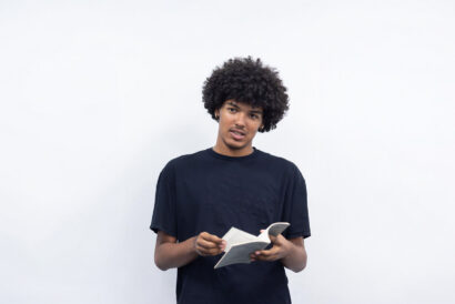 College Audition Monologue Prep Grades 10-12 - Hanover Theatre and Conservatory Young man with curly hair in a dark T‑shirt holds an open booklet and looks at the camera against a light background.