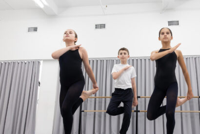 Contemporary Foundations SUMMER - Hanover Theatre and Conservatory Three young ballet dancers are mid-air in a studio, wearing black leotards (two girls) and a white T-shirt with dark pants (boy), with a barre and grey curtains in the background.