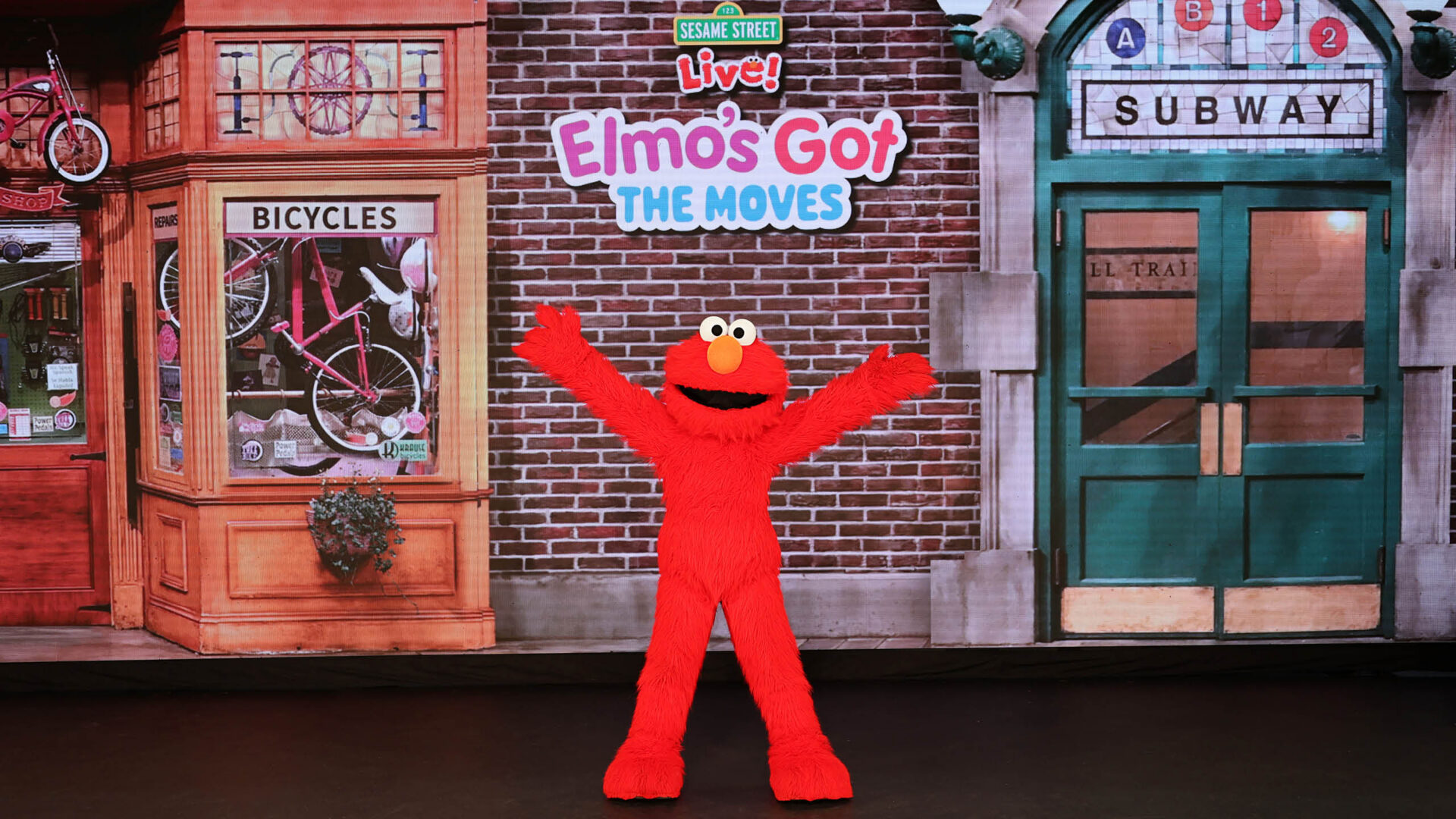 Elmo stands excitedly with his arms wide open in front of a bicycle shop and an entrance to the subway.