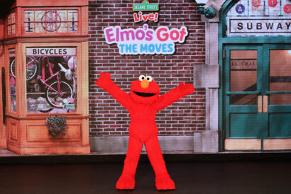 Elmo Got Moves - Hanover Theatre and Conservatory Elmo stands excitedly with his arms wide open in front of a bicycle shop and an entrance to the subway.