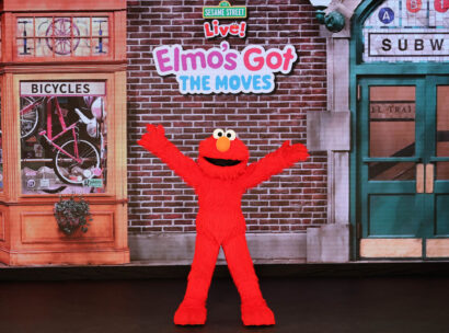 Elmo stands excitedly with his arms wide open in front of a bicycle shop and an entrance to the subway.