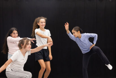 Lets Make A Musical Grades 5-7 - Hanover Theatre and Conservatory Group of young girls dancing on a stage with a black backdrop, mid-movement and smiling at the camera.
