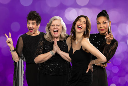 Four women in black outfits are laughing and posing together against a sparkly purple background.