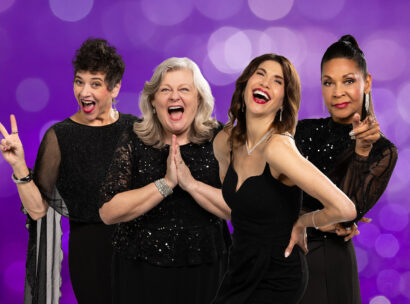 Four women in black outfits are laughing and posing together against a sparkly purple background.
