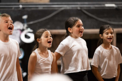 Musical Note Ninjas Grades 2-5 - Hanover Theatre and Conservatory Group of children singing together on stage, wearing white shirts during a rehearsal/break performance.