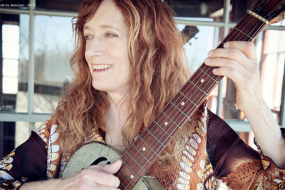 PattyLarkin_1920 x 1080 hero image - DCM - video card - FB event - Hanover Theatre and Conservatory A woman smiling playing her guitar looking off camera in front of a reflective window