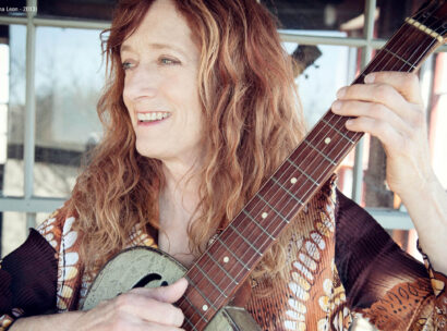 A woman smiling playing her guitar looking off camera in front of a reflective window