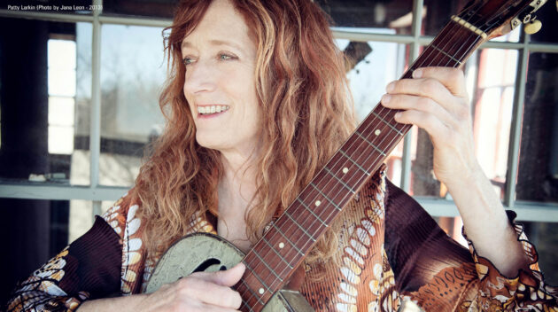A woman smiling playing her guitar looking off camera in front of a reflective window