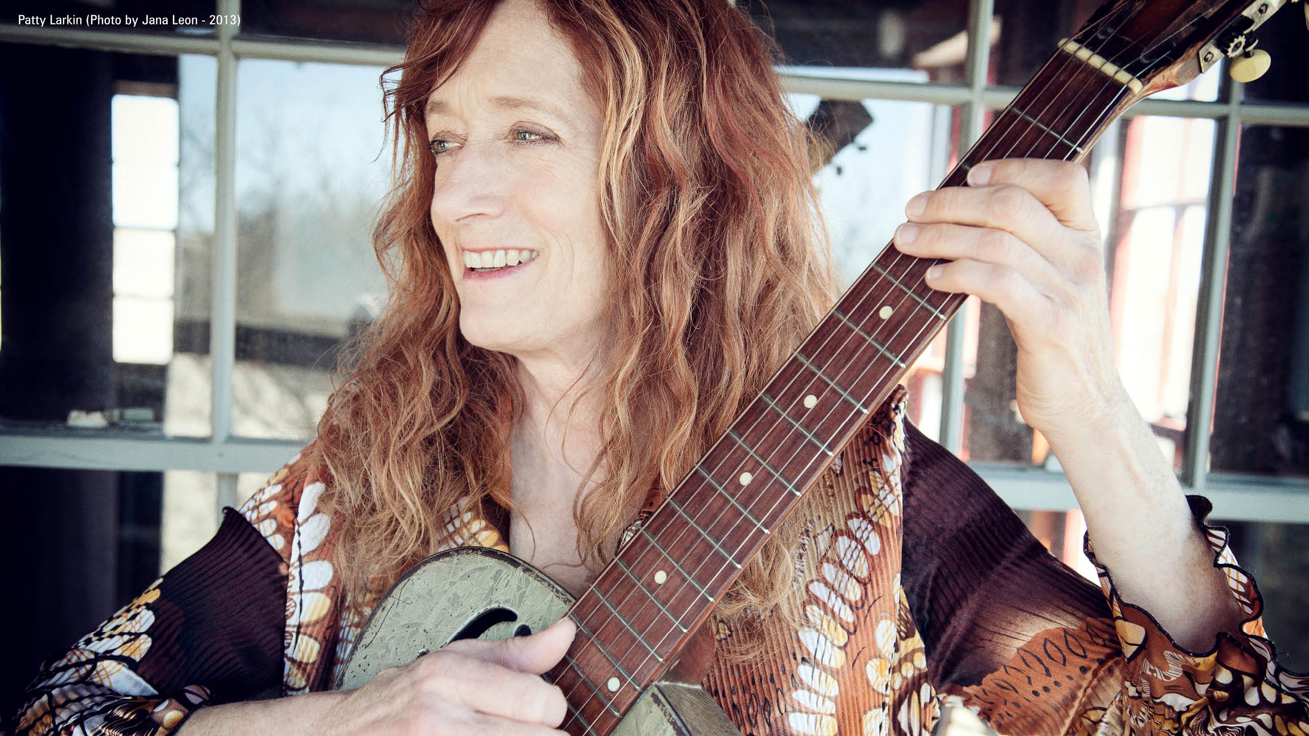 A woman smiling playing her guitar looking off camera in front of a reflective window