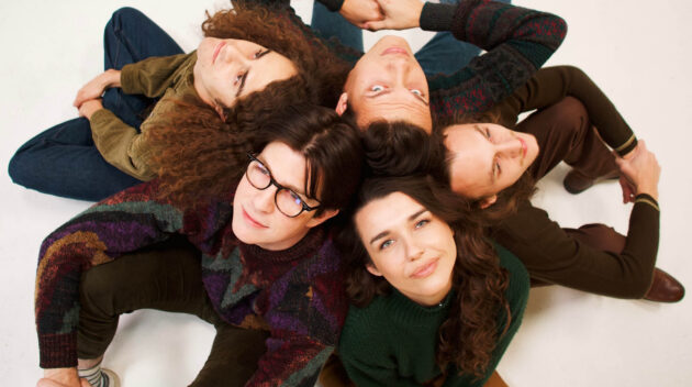 Five people sitting in a circle with their backs all touching with one woman looking at the camera