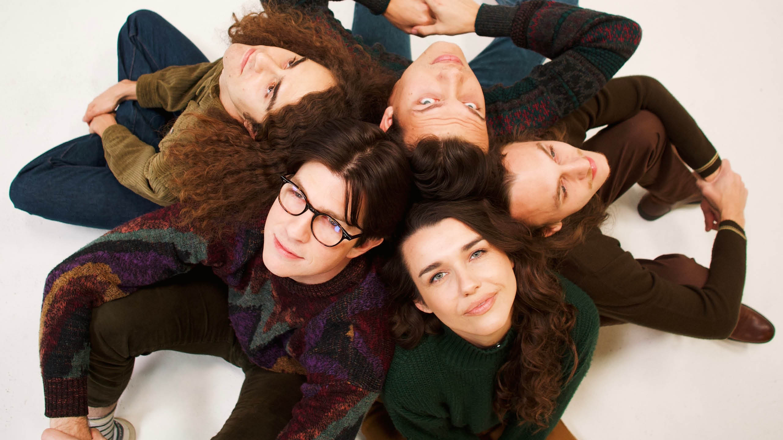 Five people sitting in a circle with their backs all touching with one woman looking at the camera
