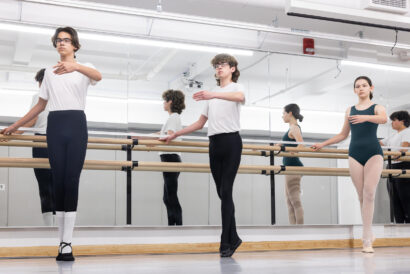 Teen Ballet Foundations SUMMER - Hanover Theatre and Conservatory Group of ballet students at a barre in a mirrored dance studio, practicing arms in first position while wearing leotards and tights.