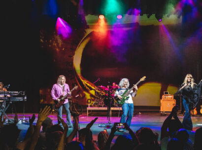 Five musicians on stage playing a song, there are bright colorful lights and a cheering crowd