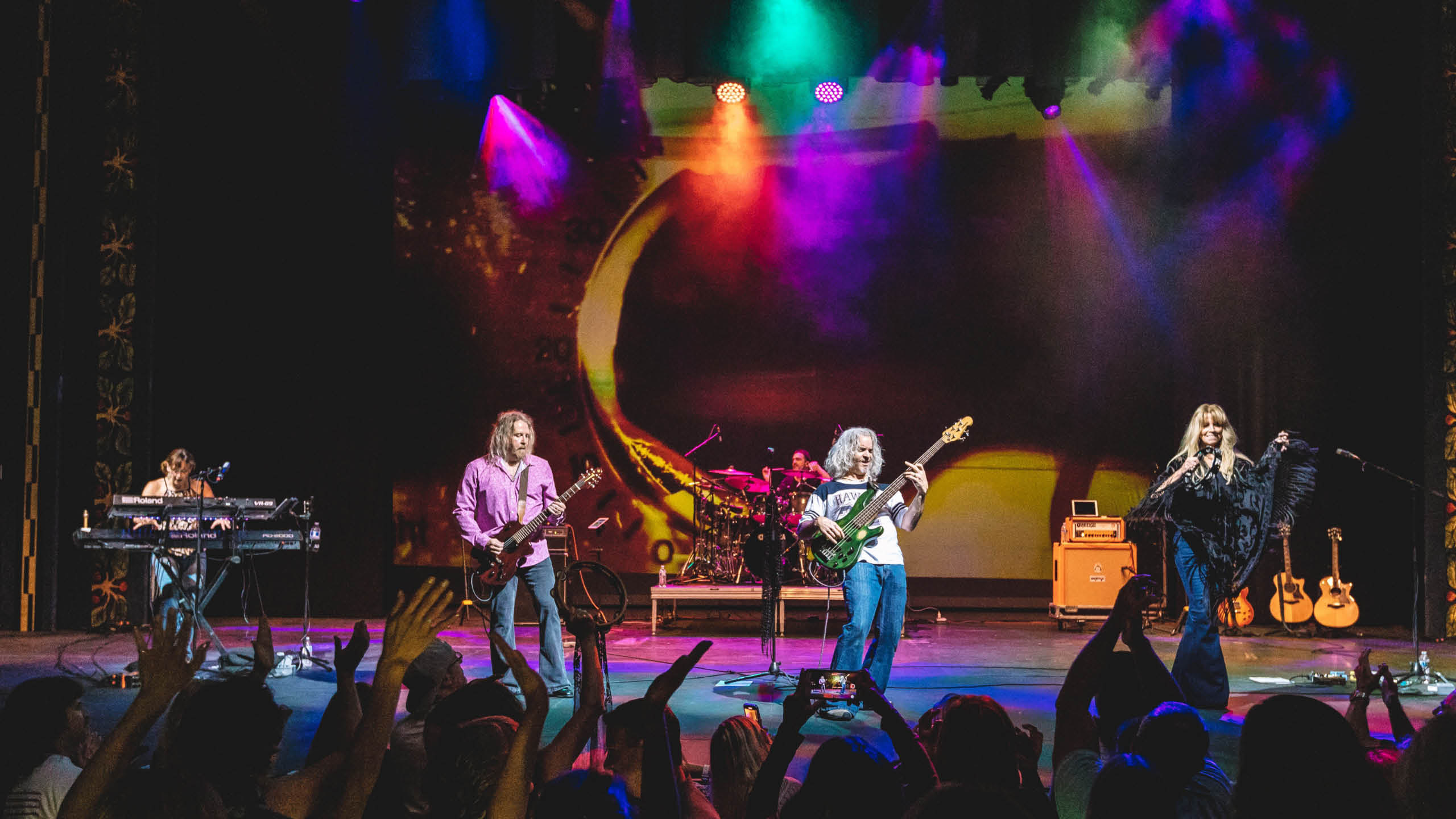 Five musicians on stage playing a song, there are bright colorful lights and a cheering crowd