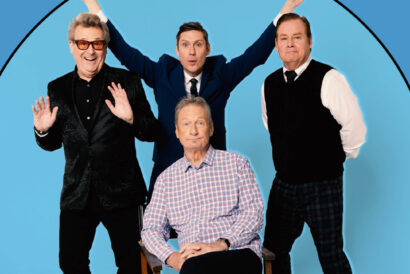 WhoseLive_NEW_1920 x 1080 hero image - DCM - video card - FB event - Hanover Theatre and Conservatory Four men posing together in a blue circular backdrop; one seated in a plaid shirt, the others standing with arms raised in celebration.