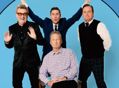 Four men posing together in a blue circular backdrop; one seated in a plaid shirt, the others standing with arms raised in celebration.