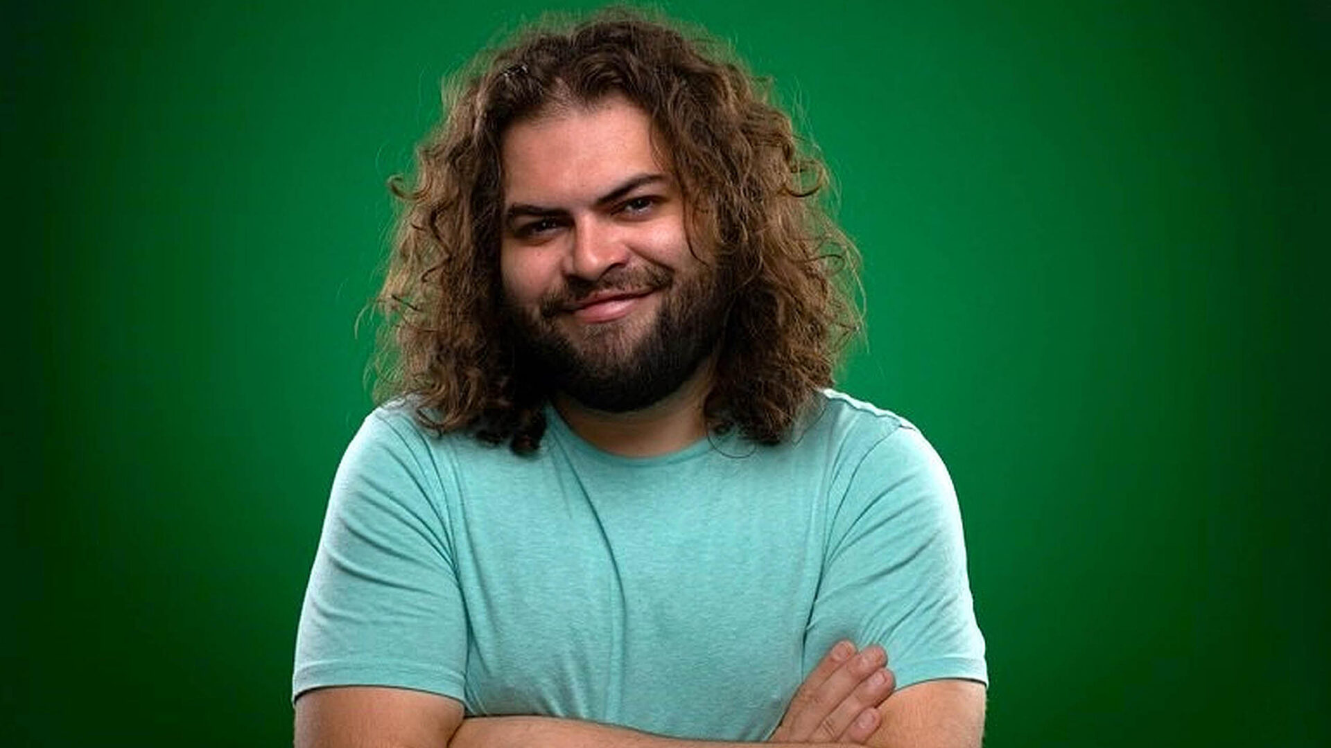 Dustin Ybarra smiles at the camera with his arms crossed wearing a blue tee shirt in front of a green background.