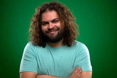 Dustin Ybarra smiles at the camera with his arms crossed wearing a blue tee shirt in front of a green background.