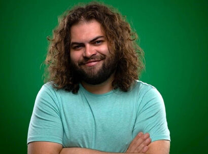 Dustin Ybarra smiles at the camera with his arms crossed wearing a blue tee shirt in front of a green background.