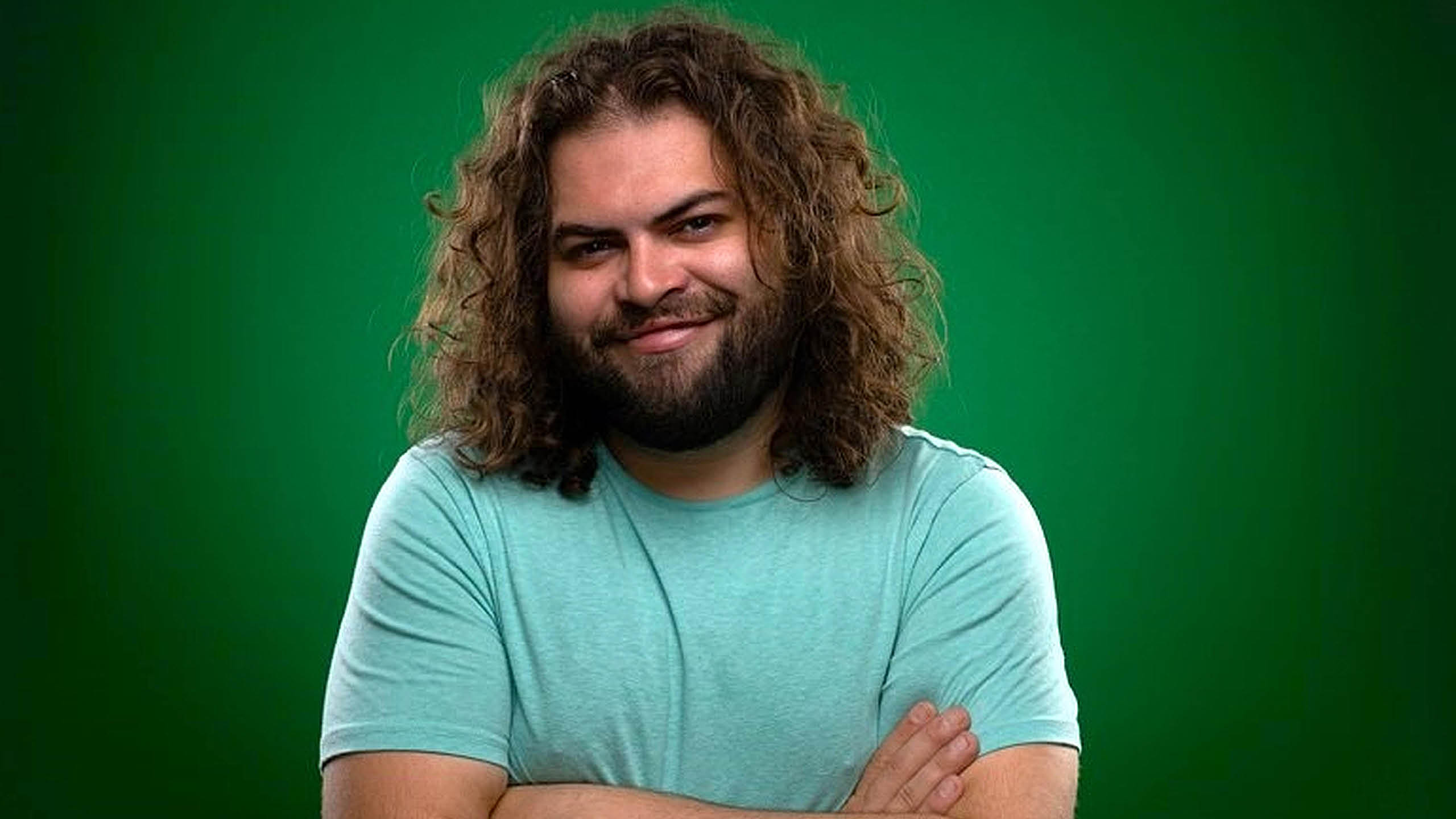 Dustin Ybarra smiles at the camera with his arms crossed wearing a blue tee shirt in front of a green background.