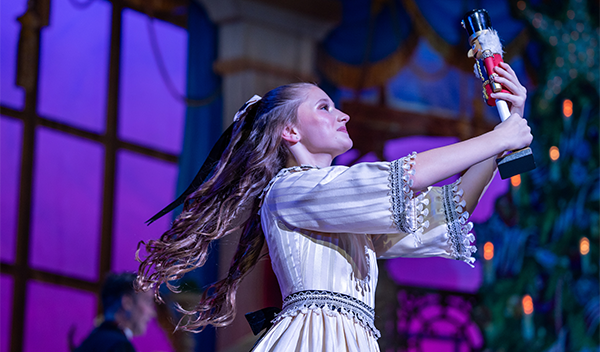 Female performer in a white costume raises a baton on a brightly lit stage.