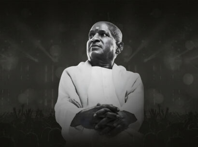 Man in white traditional robes stands with folded hands on a dark stage, a cheering crowd silhouettes at the bottom.