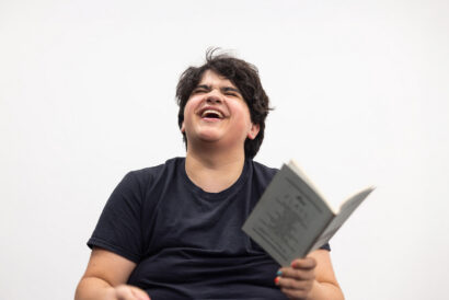 Acting 2 Scenework Grades 9-11 - Hanover Theatre and Conservatory Person with short dark hair in a black T‑shirt laughing while reading an open book against a white background.