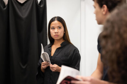 Acting 2 Scenework Grades 9-11 Option 2 - Hanover Theatre and Conservatory Woman in a black shirt holding a program, standing by a black curtain and looking to the side as if in an audition or rehearsal.