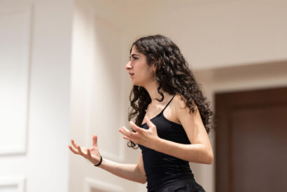 Acting 3 Classical Grades 10-12 - Hanover Theatre and Conservatory Woman with long curly hair in a black tank top speaks and uses expressive hand gestures during a presentation indoors.