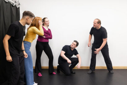 Adult Acting 2 Scenework Ages 18+ - Hanover Theatre and Conservatory Group of five adults in a studio, some standing and one kneeling, reacting playfully to a man on the right in a black shirt.