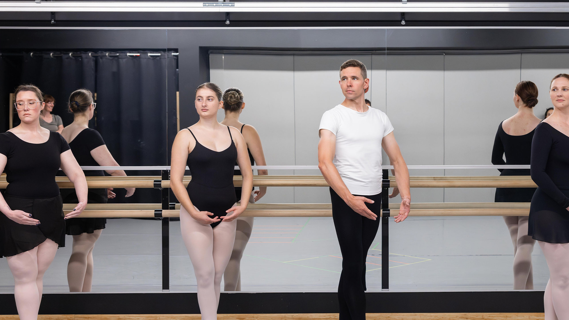 Dancers at a ballet barre in a studio, facing forward with a male in a white T-shirt and others in black leotards by a mirrored wall.