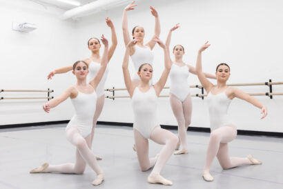 Apprentice Youth Ballet Company Invitation Only - Hanover Theatre and Conservatory Group of ballet dancers in white leotards and tights pose in a bright studio with a barre running along the wall behind them, arms raised in graceful lines.