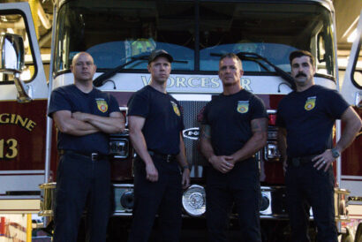 BDTH_1920 x 1080 hero image - video card - FB event - Hanover Theatre and Conservatory Four firefighters stand with arms crossed in front of a red fire engine inside a station, wearing dark uniforms and badges.