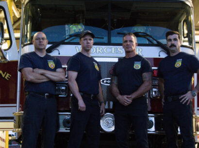 Four firefighters stand with arms crossed in front of a red fire engine inside a station, wearing dark uniforms and badges.