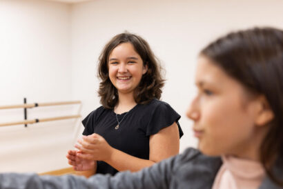 Beginner Improv 2 and Performance Grades 7-8 - Hanover Theatre and Conservatory Young woman in a black shirt smiles and claps in a dance studio with a ballet barre in the background