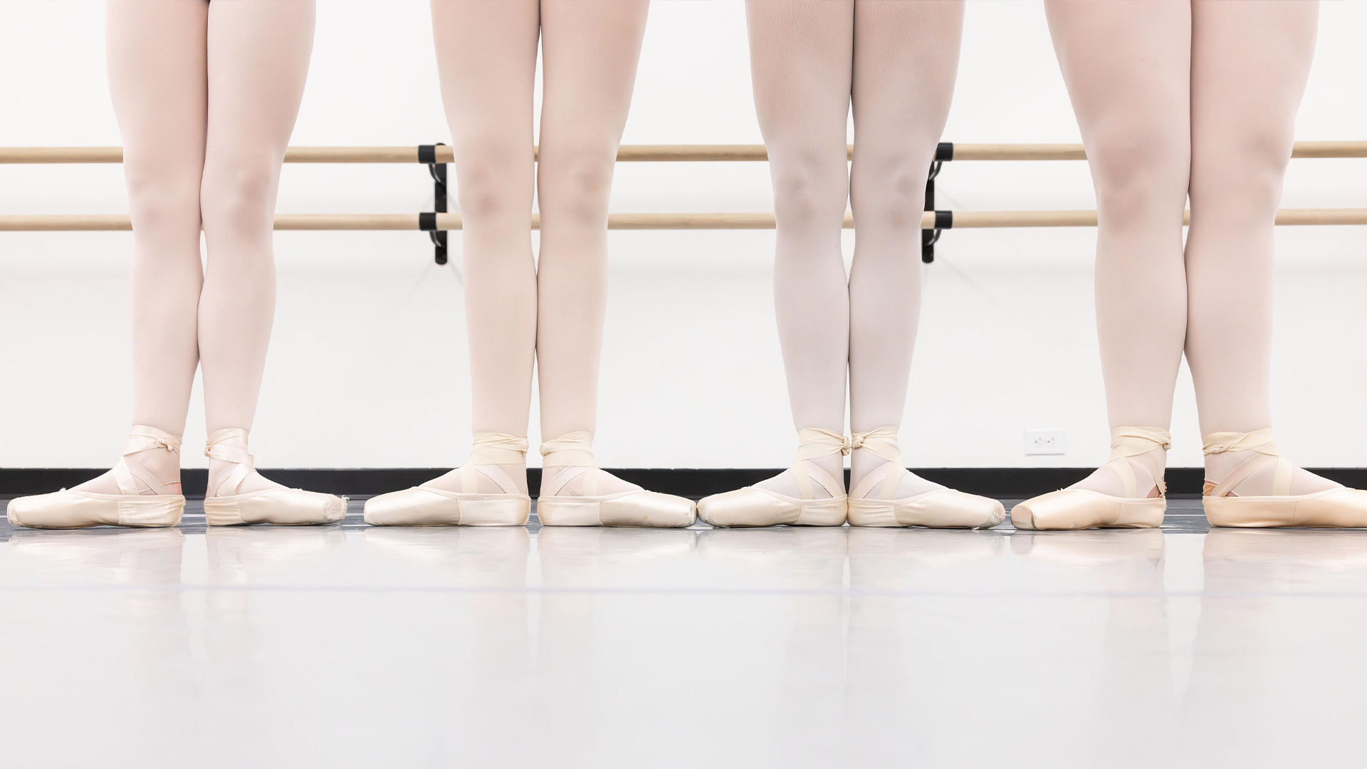 Five ballet dancers standing en pointe at a barre in pale tights.