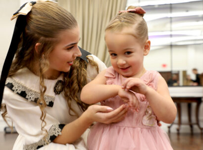 Clara, a young girl dressed in a white dress with lace details and a bow in her hair, talking to a toddler in a pink dress during Cookies and Tea.