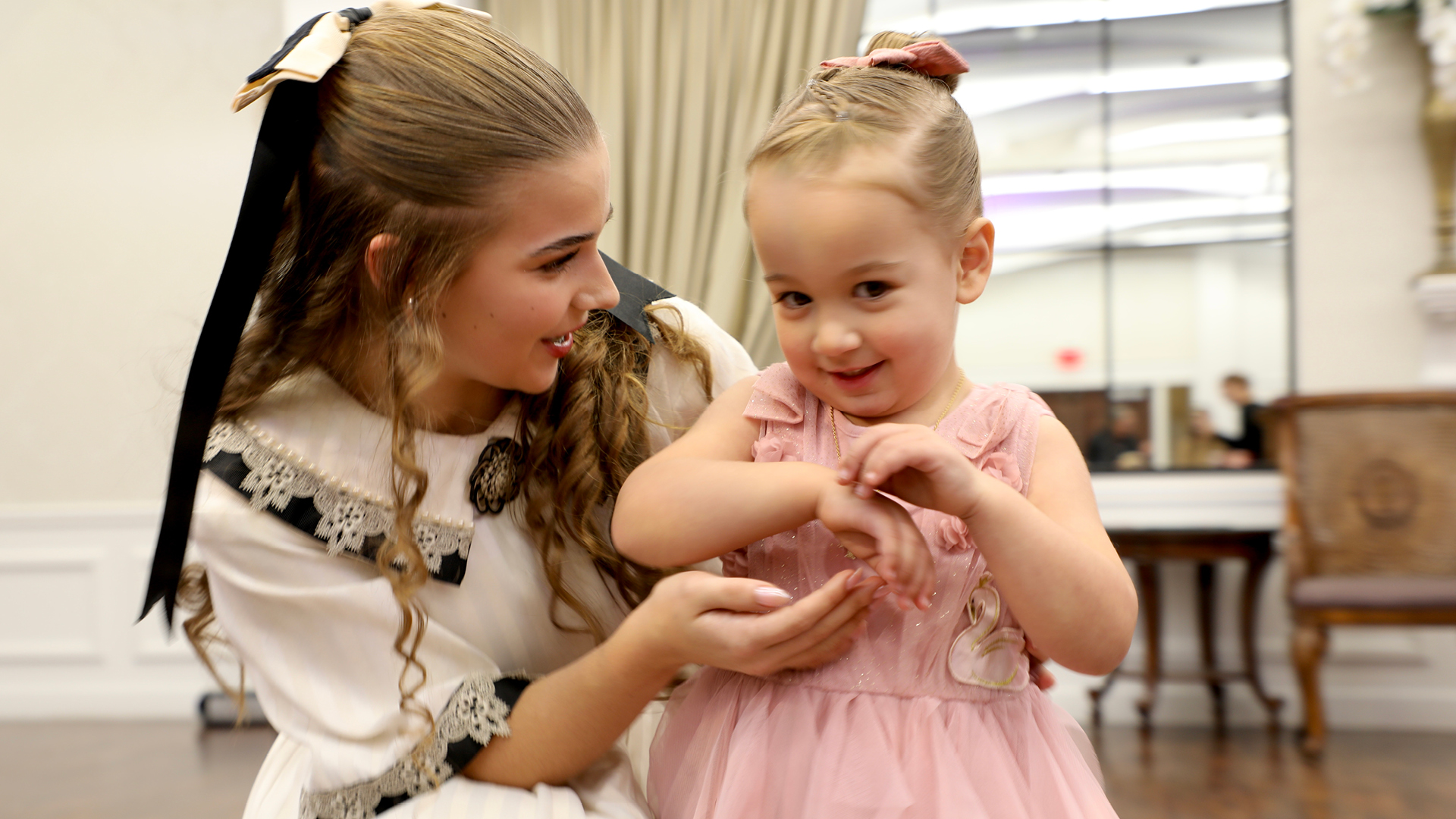 Clara, a young girl dressed in a white dress with lace details and a bow in her hair, talking to a toddler in a pink dress during Cookies and Tea.