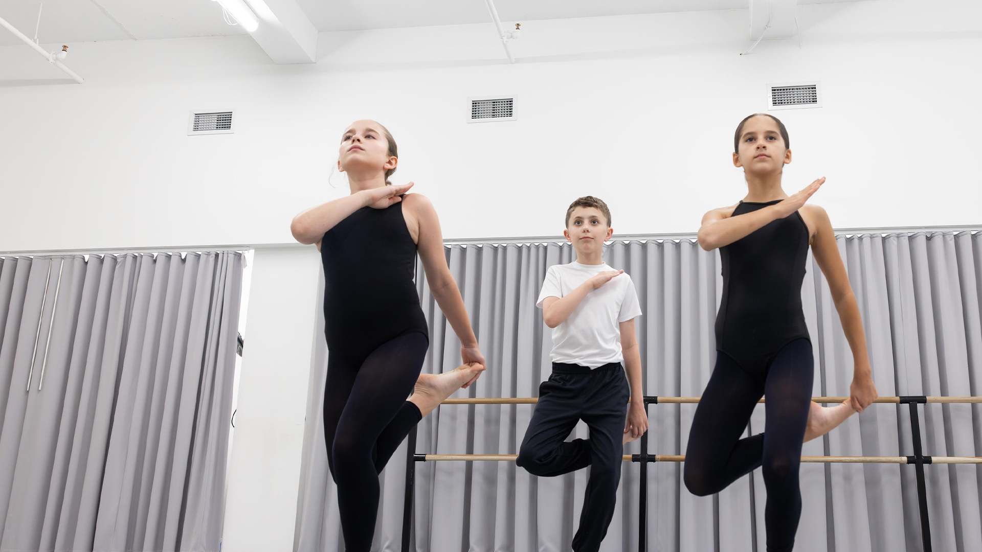 Three young ballet students leap at a barre in a bright dance studio.