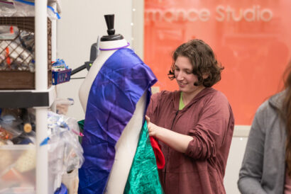 Costume Design Teens Adults - Hanover Theatre and Conservatory Woman in a maroon hoodie adjusts colorful fabric on a dress form in a workshop, with supplies on shelves nearby and an orange wall in the background.”