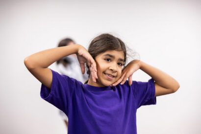 Creative Drama 1 Grades 4-5 - Hanover Theatre and Conservatory Girl in a purple shirt smiling while posing with hands near her face in a dance pose, studio background behind her