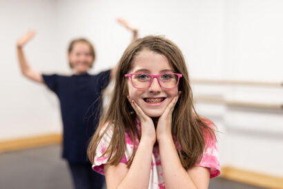 Creative Drama 1 and Performance Grades 4-5 - Hanover Theatre and Conservatory Smiling girl with braces and pink glasses, hands cupping her face, in focus; a blurred person with arms raised in the background.