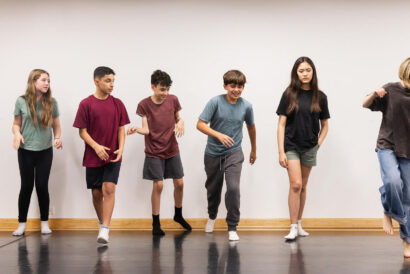 Creative Drama 2 and Performance Grades 6-7 - Hanover Theatre and Conservatory Six kids dancing in a studio with a white wall and wooden floor, wearing casual t-shirts and shorts, mid-movement.